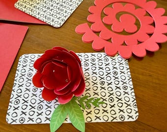 May include: A red paper flower with green leaves attached to a rectangular card featuring a black and white XOXO pattern. Nearby, a flat red spiral paper cutout and a red envelope are visible, showcasing components for a craft project.
