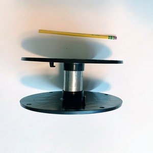 May include: A black plastic spool with a central silver metal cylinder, positioned horizontally. A yellow pencil is placed above the spool for scale, measuring approximately 7.5 inches in length, providing a clear visual reference for the size.