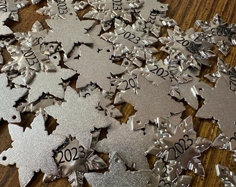 May include: A collection of silver-colored metal snowflake ornaments, each featuring a six-pointed design with intricate cutouts and the year 2023 clearly stamped in black ink on one of the points. Each snowflake includes a small hole for hanging.