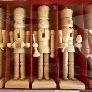 May include: A set of five wooden musical nutcrackers in a red box. Each nutcracker is light brown and has a unique design, including a drummer, a horn player, and a musician with a spoon-like instrument. The box has a label that says "MUSICAL NUTCRACKERS".