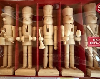 May include: A set of five unfinished light wood nutcracker figures in a red box, each holding a different musical instrument including a drum, cymbals, a trumpet, and a guitar. The packaging is labeled 5 PC and MUSICAL NUTCRACKERS.