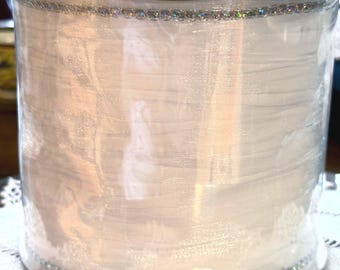 May include: A large spool of white sheer ribbon wrapped in clear plastic packaging. The ribbon features a horizontal crinkled texture and is bordered on the top and bottom edges by a thin, sparkling silver trim with small reflective rhinestones.