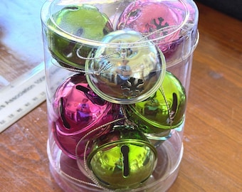 May include: A clear cylindrical plastic container filled with multiple metallic jingle bells in shades of lime green and magenta. Each bell features a classic slit opening and a small silver hanging string, with one bell displaying a snowflake cutout.