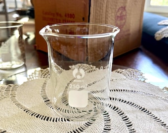 May include: A clear glass laboratory beaker with a pouring spout and a 250 ml capacity marking printed in white on the side. The beaker features a white rectangular area for labeling and a logo with the text Bomex visible on the glass surface.