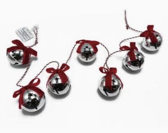 May include: A garland featuring six shiny silver jingle bells, each adorned with a small red velvet ribbon bow. The bells are connected by a red and white twisted cord, creating a festive string of ornaments for seasonal decoration.