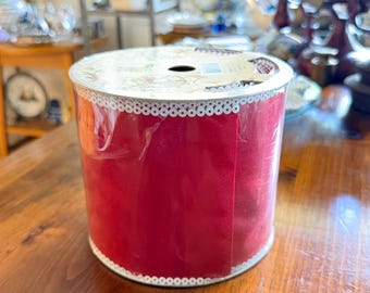 May include: A cylindrical spool of deep red ribbon wrapped in clear plastic. The ribbon features a white scalloped edge trim along the top and bottom borders. The spool has a circular cardboard top with a central hole and visible printed text.