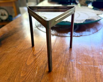 May include: A metallic, triangular-shaped laboratory tripod stand featuring three vertical legs and a flat, open-frame top. The object is constructed from a single piece of bent metal with a smooth, slightly weathered finish and a uniform structure.
