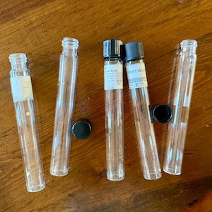 May include: Five clear glass test tubes arranged in a row. Two tubes feature black screw-on caps, while three are open. Two of the tubes have white paper labels with printed text attached to their upper sections. One loose black cap lies nearby.