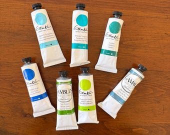 May include: Seven tubes of Gamblin Artists' Oil Color paint in various shades of blue, green, and gray. The tubes are white with black caps and feature color swatches and text labels. The tubes are arranged on a wooden surface.