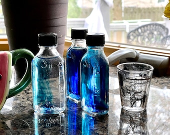 May include: Three clear glass bottles with black screw-top lids, each featuring embossed measurement markings up to 3 ounces and 40 milliliters, containing varying shades of blue liquid, alongside a small glass featuring a black graphic of a longhorn.