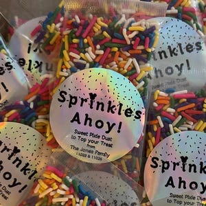 May include: Several small, clear plastic bags filled with colourful sprinkles. Each bag has a round, holographic sticker that reads "Sprinkles Ahoy!" in a playful font. The sprinkles are a mix of red, yellow, blue, and green.