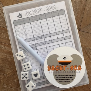 May include: A clear plastic case containing a Yacht-Sea game set. The set includes a score sheet, dice, a pen, and a round coaster with the game's logo and text. The dice have black Mickey Mouse icons.