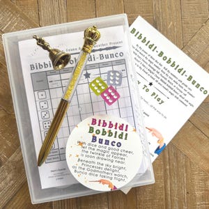 Peut inclure: Un coffret en plastique transparent contient un jeu avec un stylo doré, des dés et des cartes de jeu imprimées. Le stylo est surmonté d'une couronne et rempli de cristaux jaunes. Le jeu s'appelle "Bibbidi-Bobbidi-Bunco."