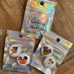 Puede incluir: Tres bolsas holográficas que contienen botones con temática de Halloween. Los botones presentan personajes de dibujos animados y calabazas. Las bolsas están etiquetadas como "Sparkling not Spooking" e incluyen el texto "Happy Halloween from The Jones Family".