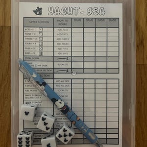 May include: A clear plastic case containing a Yacht-Sea game set. The set includes a score sheet, dice, and a Mickey Mouse-themed pen. The dice are white with black Mickey Mouse icons. The score sheet has the game's title and scoring sections.