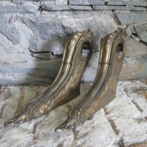 May include: Two ornate gold-colored metal brackets with decorative scrollwork and paw-like feet. The brackets are lying on a white fur surface.