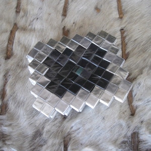 May include: A square, geometric coaster composed of small, reflective silver and black squares. The coaster has a diamond pattern and is displayed on a textured, light-colored surface.