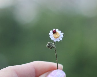 Puede incluir: Una pequeña margarita de ganchillo con una mariquita roja en la parte superior, sostenida en una mano contra un fondo verde borroso.