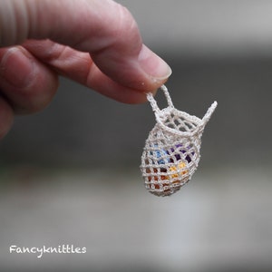 Può includere: Una piccola borsa bianca all'uncinetto con chiusura a cordoncino, piena di biglie colorate. La borsa è tenuta da un dito.