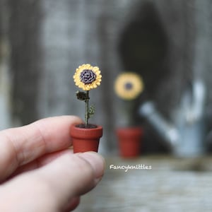 May include: A miniature crocheted sunflower plant in a terracotta pot. The sunflower is yellow with a brown centre. The plant is being held in a hand.