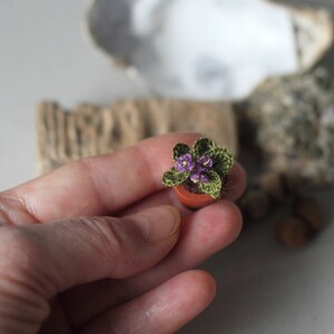 Peut inclure: Une miniature de violette africaine crochetée dans un petit pot en terre cuite. La plante présente des feuilles vertes et de petites fleurs violettes. L'article fait main est une œuvre d'art délicate.