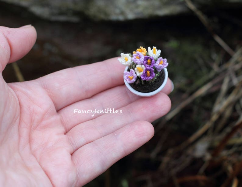 May include: A small white pot with a cluster of purple and white crocheted crocuses. The pot is sitting in a hand.