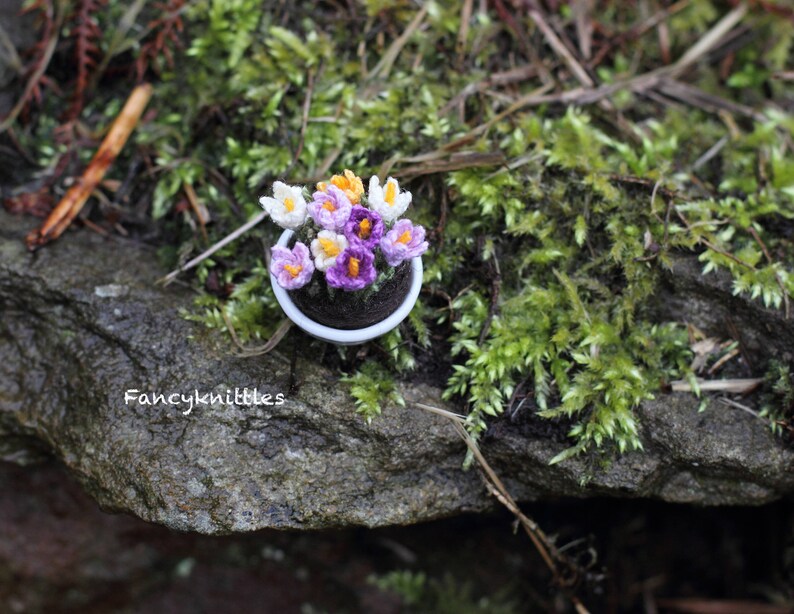 May include: A small white pot with purple and yellow crocheted crocuses. The pot is sitting on a mossy rock.