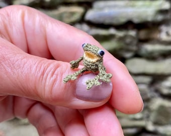 Op de afbeelding: Een kleine gehaakte kikker van groen en bruin garen, zittend op een hand met zijn mond open en ogen omhoog gericht.