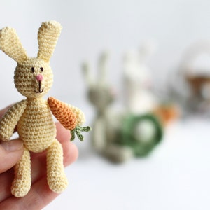 May include: A small crocheted yellow bunny rabbit holding a small orange carrot in its right hand. The bunny is sitting on a hand.