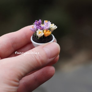 May include: A small white pot with a crocheted bouquet of purple, white, and orange crocuses. The pot is sitting in a hand.