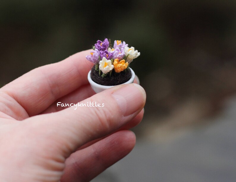May include: A small white pot with a crocheted bouquet of purple, white, and orange crocuses. The pot is sitting in a hand.