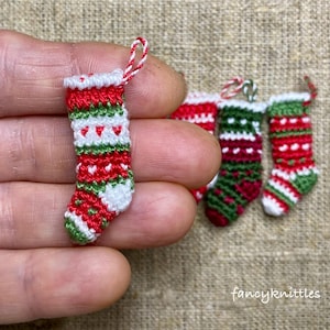 Könnte beinhalten: Miniatur-Weihnachtsstrümpfe in Rot, Grün und Weiß mit Herz- und Streifenmustern. Ein Strumpf wird in einer Hand gehalten, weitere im Hintergrund. Die Strümpfe haben eine Schlaufe zum Aufhängen. Der Text "fancyknittles" ist sichtbar.