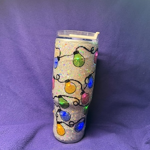 May include: A cylindrical tumbler with a silver glitter finish featuring a pattern of black wire lines and colorful light bulb illustrations in shades of blue, green, yellow, and pink. The tumbler includes a clear plastic lid with a sliding tab.