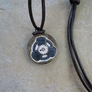 May include: A brown leather necklace with a round pendant featuring a black and white design of a gear with three spokes.