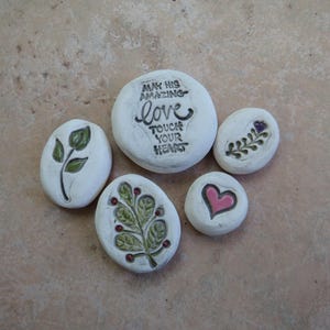 May include: Five white painted stones with various designs. One stone has the text "MAY HIS AMAZING LOVE TOUCH YOUR HEART". Other stones feature a leaf, a sprig, and a pink heart. The stones are arranged on a light brown surface.