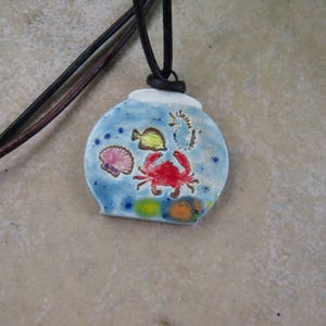 May include: A blue ceramic pendant necklace with a black cord. The pendant is shaped like a fishbowl and features a red crab, a yellow fish, a white seahorse, and a pink coral.