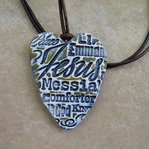 May include: A heart-shaped pendant necklace with a brown leather cord. The pendant is white with gold and black lettering that reads "Peace, Emmanuel, Jesus, Messiah, Comforter, The God King".