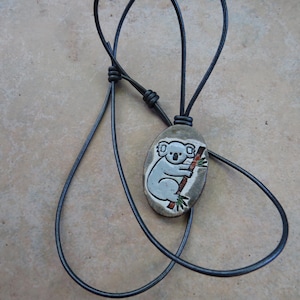May include: A black leather cord necklace with a ceramic pendant featuring a cartoon koala bear sitting on a tree branch. The pendant is oval-shaped and has a white background with brown and green details.