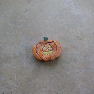 May include: A ceramic pumpkin with a carved face, painted in shades of orange, brown, and yellow. The pumpkin has a green stem and is likely a Halloween decoration. The face has triangular eyes and a jagged mouth.