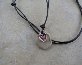 May include: A handmade necklace featuring a natural stone pendant with a small, heart-shaped ceramic accent. The ceramic has a white border and a pink heart design. The necklace has a dark brown leather cord.