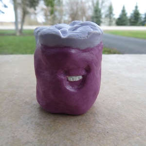 May include: A purple ceramic figurine with a smiling face and a light purple top. The figurine is shaped like a jar or container.