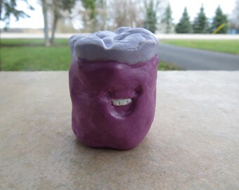 May include: A purple ceramic figurine with a smiling face and a light purple top. The figurine is shaped like a jar or container.