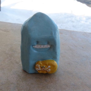 May include: A light blue ceramic figurine shaped like a house with a smiling face and a yellow cat figurine attached to the front.