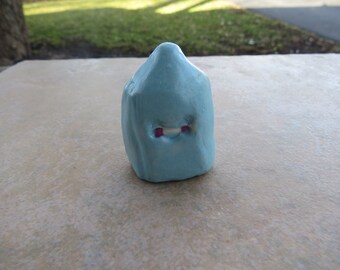 May include: A small, light blue ceramic sculpture shaped like a house with two small pink eyes.