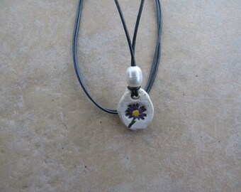 May include: A white ceramic pendant necklace with a purple and yellow flower design. The necklace is on a black cord with a white pearl bead.