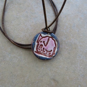 May include: A brown leather cord necklace with a round ceramic pendant featuring a pink elephant design. The pendant has a blue and brown glaze.