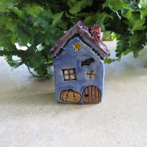 May include: A small ceramic house figurine featuring a blue facade, a purple roof with a red chimney, and a yellow star detail. The front displays a painted orange pumpkin, a brown arched door, and two windows with white panes and black frames.