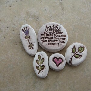 May include: Five white, oval-shaped stones with various designs. One stone has the text "faith IS BEING SURE OF WHAT WE HOPE FOR AND CERTAIN OF WHAT WE DO NOT SEE HEB 11:1". Other stones feature a purple flower, a pink heart, and green leaves.