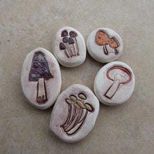 May include: Six ceramic mushroom-shaped coasters with different designs. The coasters are white with brown, orange, red, and purple painted details.