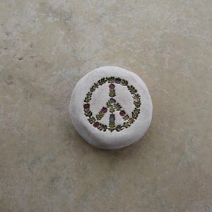 May include: A round, off-white stone with a peace symbol design. The peace symbol is made of a wreath of green leaves and purple and red flowers. The stone is on a light brown surface.